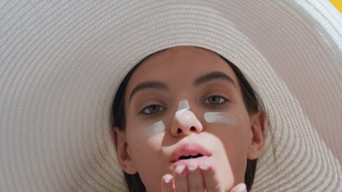 Close-up beauty portrait of young brunette Caucasian woman in a big white hat with sunscreen on her cheeks and nose blows a kiss for the camera against yellow background | Skin care commercial concept