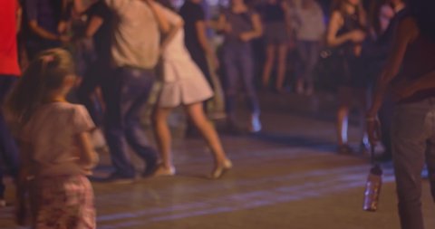 Many couples dancing latin-american dances outside little girl trying to do the same dance moves in foreground. Street salsa dance in the night, many young couples dance social Latino dances defocused