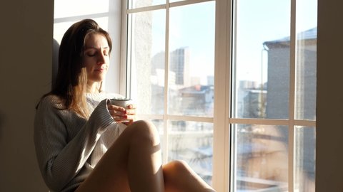 Woman is drinking a cup of hot tea, cocoa with marshmallows and relaxing at home, sitting by the window. Taking care of health, authenticity, a sense of balance and tranquility. The concept of balance