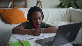 Online education at home. spbd Concentrated African-American boy pupil with headphones writes in copybook watching videolesson via laptop indoors