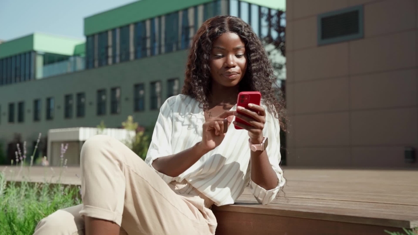 Young African black happy woman holding smartphone, surfing, using mobile apps for online shopping, e learning, dating or social media applications on cell phone sitting outdoor in modern city. | Shutterstock HD Video #1082465566
