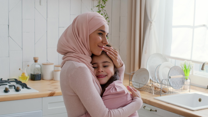 Muslim Mommy In Hijab Hugging And Kissing Little Daughter Expressing Her Love Sitting In Modern Kitchen At Home. Happy Middle Eastern Mother And Her Child Bonding Indoor. Slowmo Royalty-Free Stock Footage #1082471680