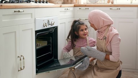 Excited Muslim Mother And Daughter Baking, Taking Cookies From Oven Cooking In Modern Kitchen At Home, Wearing Gloves. Middle Eastern Family Making Pastry Together On Weekend