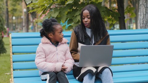 Young african american mom with daughter on bench in city park friendly family together looking in laptop outdoors cute kid black little girl confidently enthusiastic conversation with smiling mother