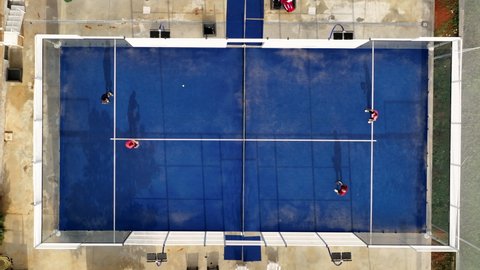 View from above, aerial footage, slow motion video of some people playing on a blue padel court. Padel is a mix between Tennis and Squash. It's usually played in doubles on an enclosed court.