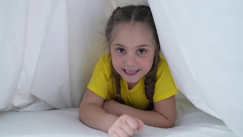 Cute little girl is crawling under covers. Baby plays on bed. Happy girl under white blanket. Children games. Smile on girl face. Portrait of a baby crawling under covers. Cute little girl play on bed