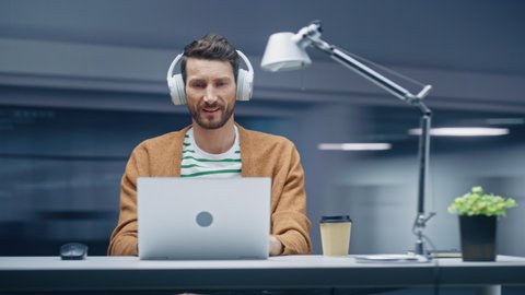 360 Degree Modern Office: Caucasian Businessman Sitting at the Desk Working on a Laptop Computer. Man Wearing Headphones, Listens to Music, Podcast. Energetic Fast Tracking Shot. Camera Moving Around