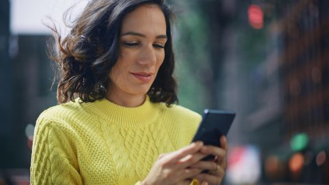 Portrait of Beautiful Happy Latin Woman Standing, Using Smartphone on a City Street. Smiling Gorgeous Hispanic Female with Lush Dark Hair using Mobile Phone for e-commerce, Online Shopping