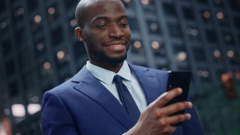 Portrait of Successful African-American Businessman using Smartphone on Street in Big City. Smiling Black Digital Entrepreneur uses Mobile Phone e-Commerce App for Stock Market Investment, Online Shop
