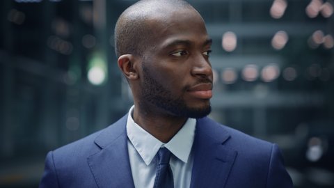 Portrait of Thoughtful Black Businessman wearing Suit, Standing in the Big City Business District Street, Turning and Looking at Camera and Smiling. Successful African American Digital Entrepreneur