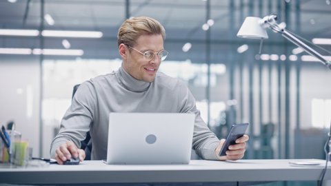Modern Office: Portrait of Caucasian Businessman Wearing Glasses Working on Laptop Computer, checks Smartphone. Digital Entrepreneur does Data Analysis for e-Commerce Startup Strategy. Static Shot