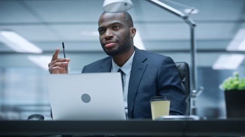 360 Degree Office: Successful Black Businessman Sitting at Desk Thinking, Using Laptop Computer. African American Entrepreneur Doing Stock Market Investment Analysis. Moving Around Tracking Shot