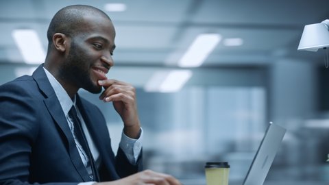 360 Degree Office: Happy Successful Black Businessman Sitting at Desk Using Laptop Computer. African American Entrepreneur in Suit working with Stock Market Investing. Moving Around Tracking Shot