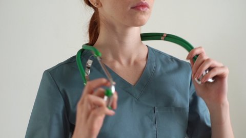Close-up cropped shot of unrecognizable female practitioner in green medical uniform putting stethoscope around neck standing on white isolated background in studio. Shooting in slow motion.
