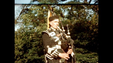 1940s: Man plays bagpipes in field. Highland men stand in field and talk. Green plaid tartan cloth hands from soldiers.