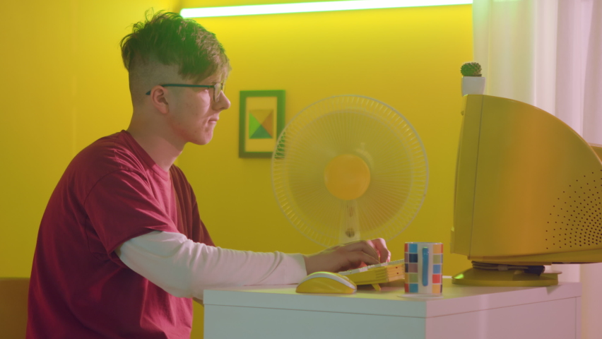 Young man, freelancer, IT specialist, is sitting in yellow office at workplace and typing on retro computer, electric fan is working in the corner, Foreground, Slow motion.