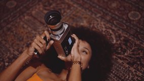 Medium Handheld Slow Motion Of Young Woman With Afro Hair In Orange Dress Using Vintage 8Mm Camera While Lying On Rug