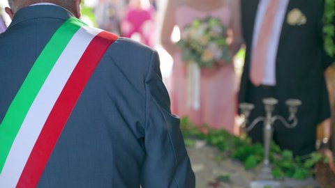 Back view of traditional Italian wedding officiant with three colours flag ribbon across the shoulder holding wedding speech in front of newlywed couple, beautiful wedding ceremony outdoor under the