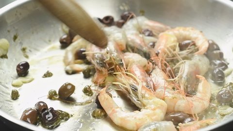 Cooking and stirring of fresh shrimps with capers and olives in a frying pan.