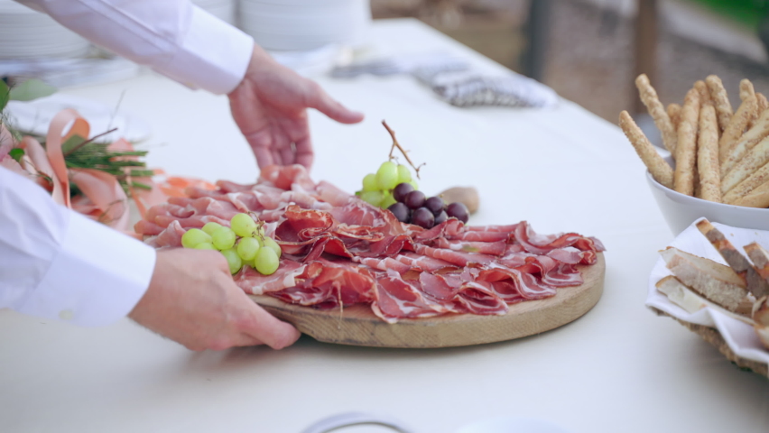 Exclusive wooden board with delicious slices of traditional Italian prosciutto and salami decorated with fruits, waiter setting banquet table with luxury restaurant menu, catering service outdoor | Shutterstock HD Video #1082499613