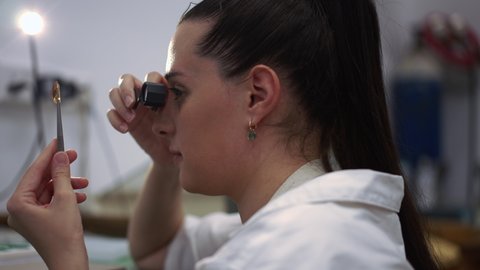Side view close-up focused female appraiser examining fire opal with loupe in jewellery workshop. Concentrated Caucasian young beautiful woman controls gemstone quality indoors. Slow motion