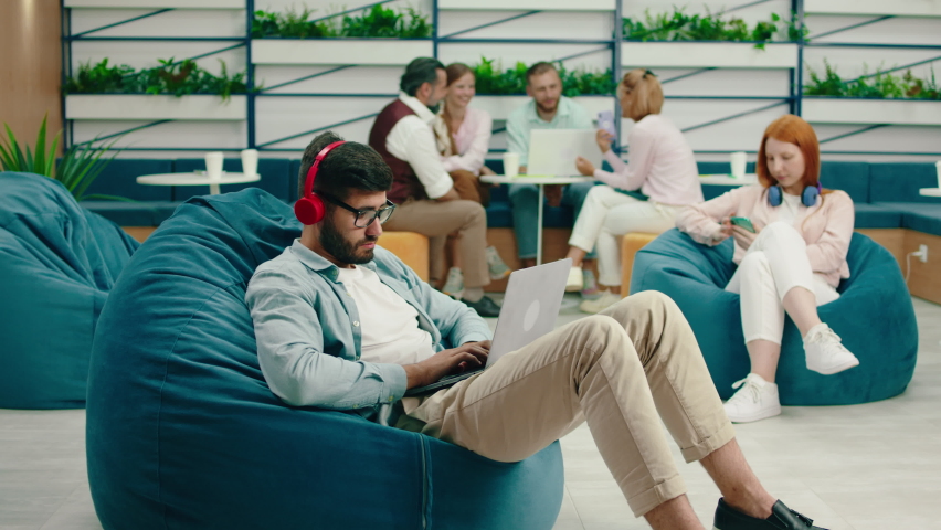 A very smart looking brunette man with a beard and glasses is doing work seriously on his laptop while listening to music on his headphone and sitting on a bean bag chair. Arri Alexa Mini. Royalty-Free Stock Footage #1082502145