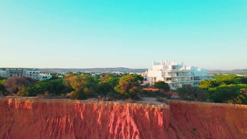 Scenic aerial view of red rocky cliffs of Praia da Falésia, Vilamoura, Algarve. Portugal sandy beach with luxury houses from above by red rocks seashore with smooth sea waves during sunset in 4K.