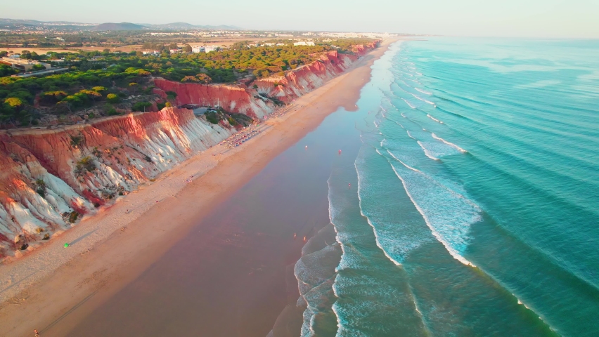 Scenic aerial view of red rocky cliffs of Praia da Falésia, Vilamoura, Algarve. Portugal sandy beach with luxury houses from above by red rocks seashore with smooth sea waves during sunset in 4K.