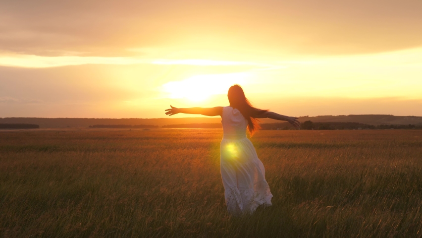 Happy young woman dancing in the spring in the sun on the field. A beautiful girl in a white dress is having fun in the summer outdoors in the rays of the sunset. Happiness female freedom concept