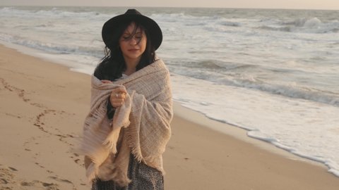 Portrait of romantic brunette woman in hat and plaid walking on the beach in autumn at sunset or sunrise 