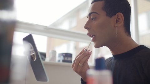 Portrait shot of androgynous male applying lip gloss in front of the mirror, in slow motion