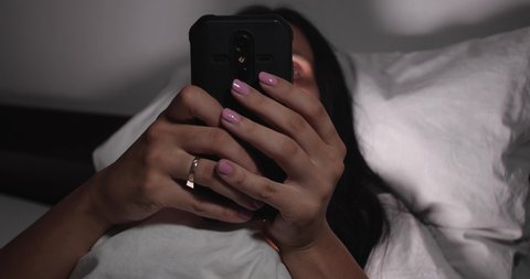 Close up of unrecognizable brunette lying, awake in bed with white linens in light room. Woman having an insomnia and looking at his cellphone. Scrolling social media or using application 4K