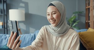 Attractive cheerful young Asia muslim beauty woman in hijab with casual sitting the couch use smartphone selfie call video conference with friend in living room at home. Girl Islam lifestyle concept.
