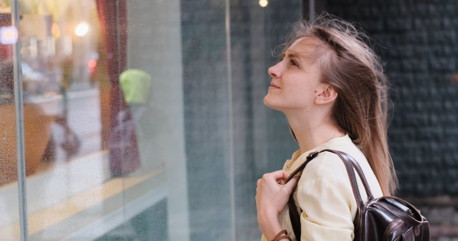 Young woman walks around the city and looks at the shop windows. Girl in casual style looks out the window of store. Wind develops hair.