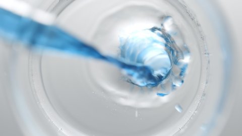 Blue liquid pouring into beaker with water creates different sized bursting bubbles and ripples on its surface on light grey background | Abstract skin nourishing cosmetics mixing concept
