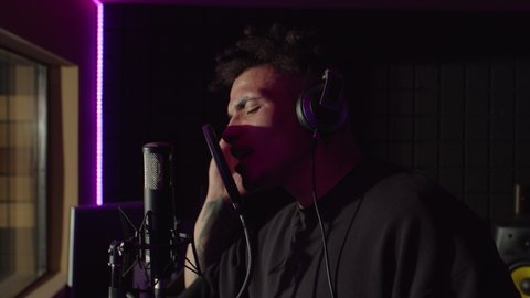 Portrait of charismatic handsome male soul singer in headphones emotionally performing new song, singing into microphone with passion while recording hit track in music studio.