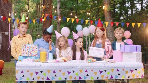 Birthday girl. Children in party caps celebrating birthday at the nature. Table with delicious food and gifts. Friends congratulating their bestie. Happy children at the birthday party concept