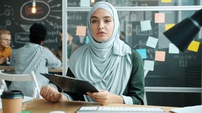 Portrait of muslim businesswoman making video call from office speaking showing charts and looking at camera. People and distant communication concept.