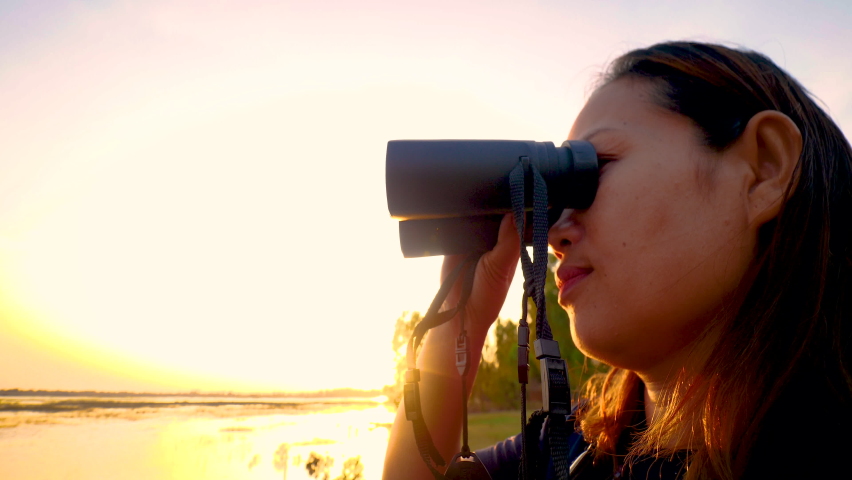 Woman traveling at reservoir looks at the view with binoculars searching for birds with sunset background.