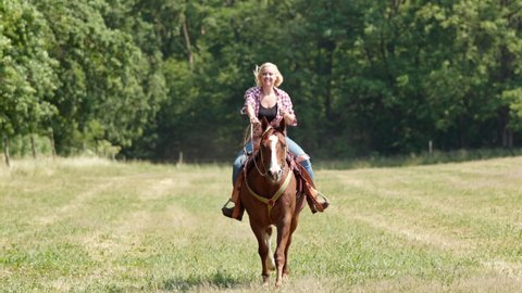 A cowgirl with blonde curls and a checked shirt rides a brown horse at a gallop towards the camera.  
