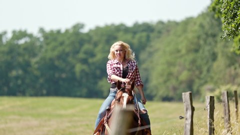 A cowgirl with blonde curls and a checked shirt rides a brown horse slowly along the edge of the forest in a field behind a fence.