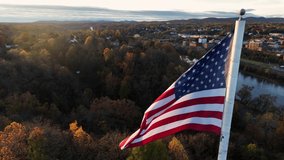 Cinematic aerial of American flag waving in breeze during autumn golden hour sunrise light. Colorful foliage, sunlight. America the Beautiful, Proud to be an America. Town reflects in river. - Powered by Shutterstock - Get 15% off with code: PIKWIZARD15