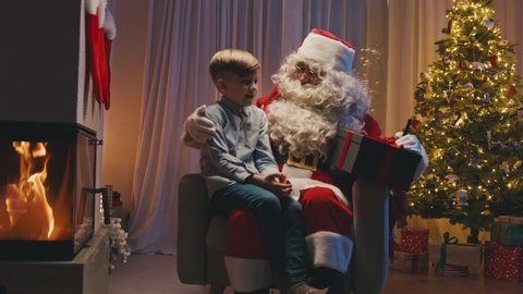 Little cute boy sitting near fireplace on Santa's knees, making a wish. Whitebearded Santa is hugging him and holding a gift.