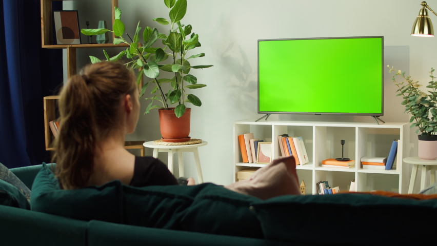 Woman watching TV with green screen in living room back view. Female person switching channels, using remote control, sitting on sofa. Domestic Cinema concept, television with chroma key.  Royalty-Free Stock Footage #1082567209