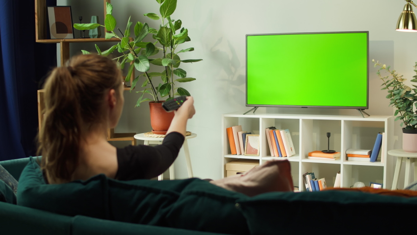Woman watching TV with green screen in living room back view. Female person switching channels, using remote control, sitting on sofa. Domestic Cinema concept, television with chroma key.  | Shutterstock HD Video #1082567209