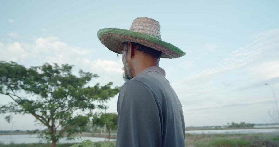 A stern bearded Asian farmer in a hat goes fishing in the evening. He turned his face and smiled slightly.
