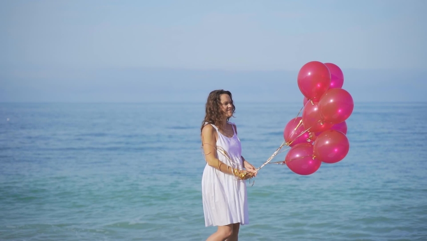 Happy Girl in white dress holds red balloons against the background of the sea and the beach. Young woman celebrates her birthday on the beach, rejoices, runs, smiles, laughs