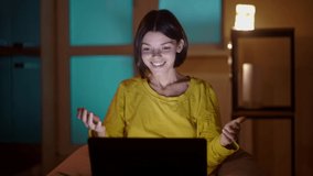 A young cute girl is surprised, feels excitement and happiness, sitting in front of a laptop in her cozy room in the evening. 