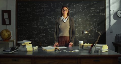 Cinematic shot of young confident female teacher or student is looking and smiling in camera in front of blackboard with math calculation,formulas and equations in college or university classroom.