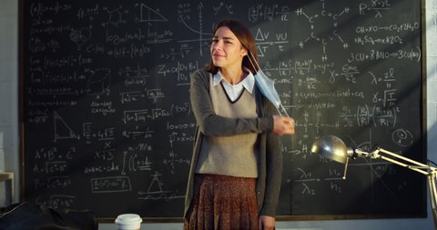 Cinematic shot of young female teacher or student is taking off protective medical mask and smiling in camera in front of blackboard with math formulas and equations in college or university classroom